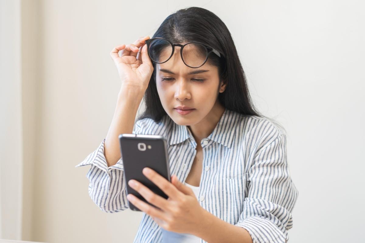 A person squinting and lifting their glasses up as they look at their phone